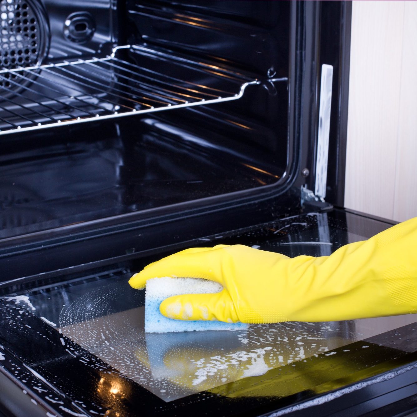 A person wearing a yellow glove is cleaning the inside of an oven with a sponge.