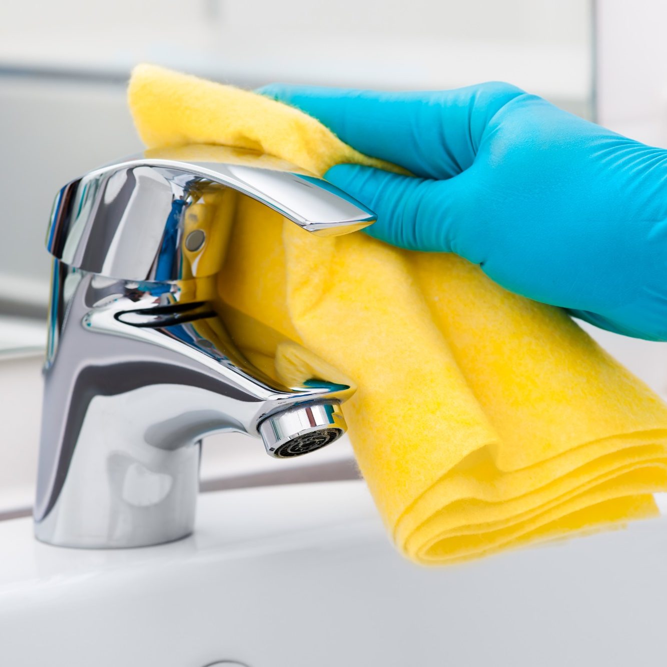 A hand with a blue glove cleaning a tap with a yellow cloth.